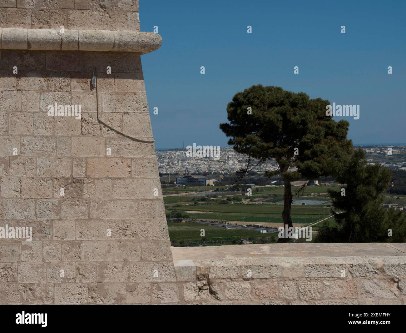 Muro di pietra in primo piano con vista su un singolo albero e un vasto paesaggio verso la lontana città, mdina, il mediterraneo, Malta Foto Stock