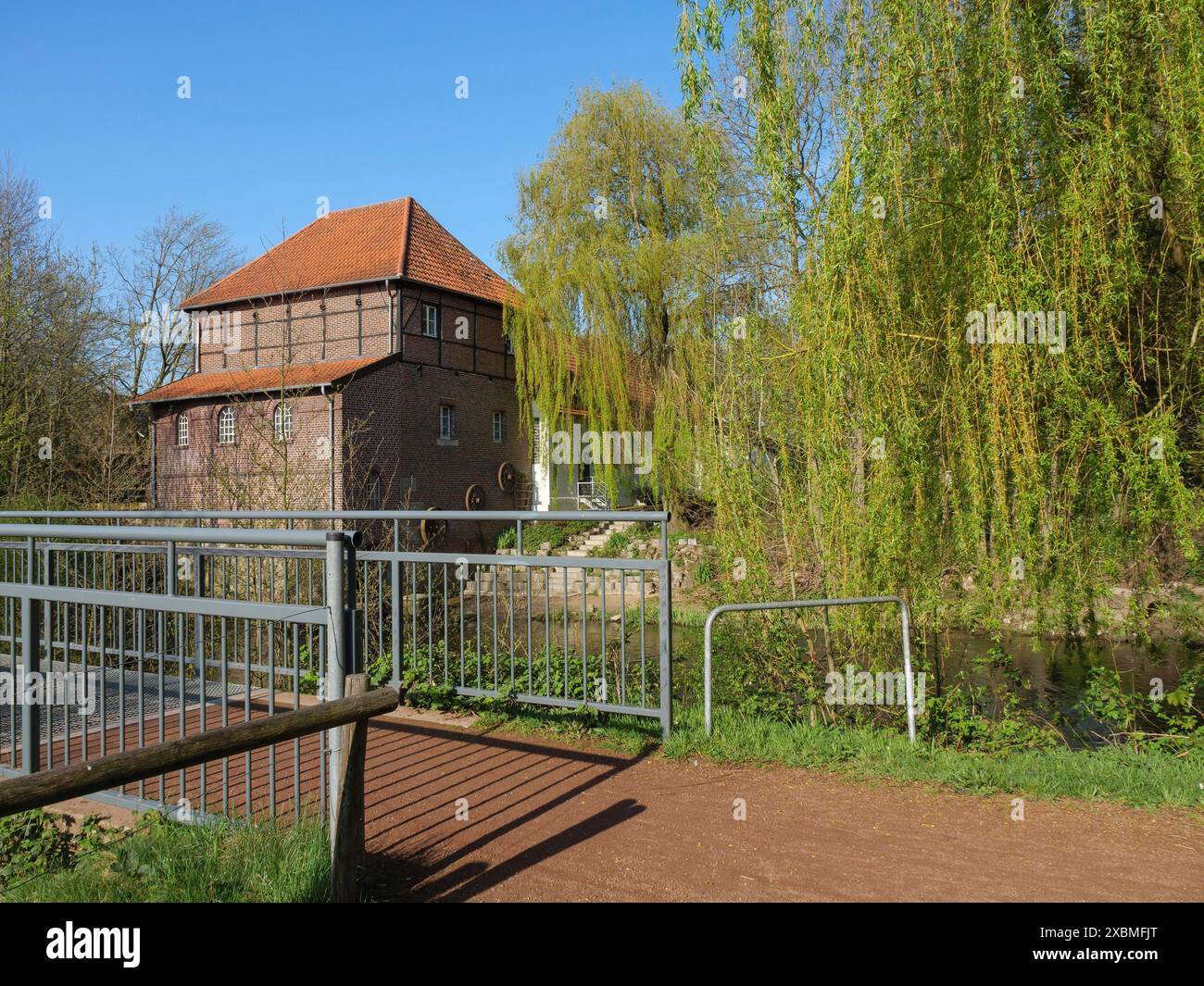 Uno stretto sentiero con un ponte e ringhiere in metallo che conduce ad un edificio in mattoni con un tetto rosso, circondato dal verde della natura e da un cielo limpido, metelen Foto Stock