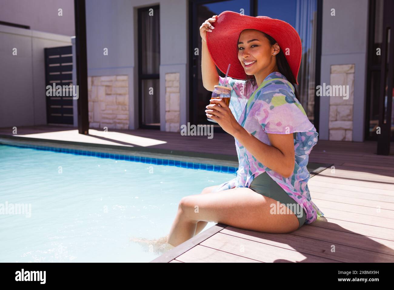 Felice donna birazziale che indossa un grande cappello rosso seduto a bordo piscina e gusta un drink Foto Stock