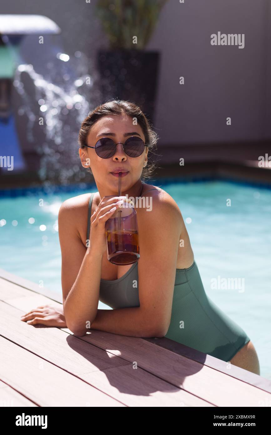 Felice donna birazziale con occhiali da sole che si rilassa a bordo piscina, sorseggiando una bevanda fresca Foto Stock