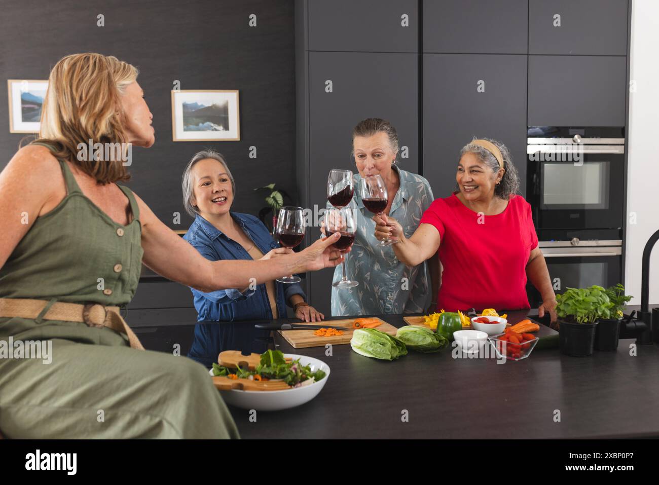 Gruppo di donne anziane che si godono il vino e preparano i pasti insieme in una cucina moderna Foto Stock