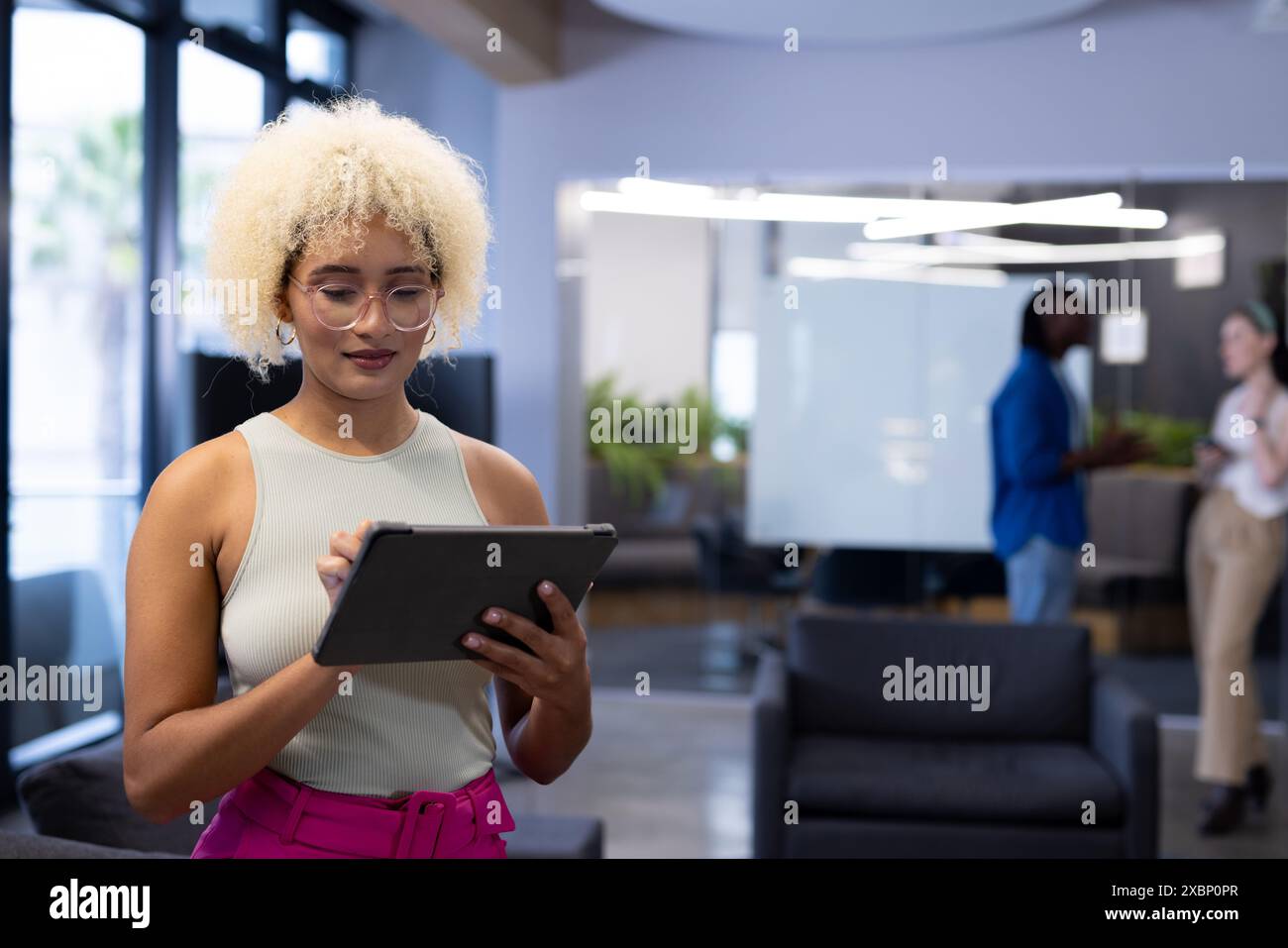 Giovane donna birazziale che usa il tablet in ufficio mentre i colleghi chiacchierano in background, copiano lo spazio Foto Stock