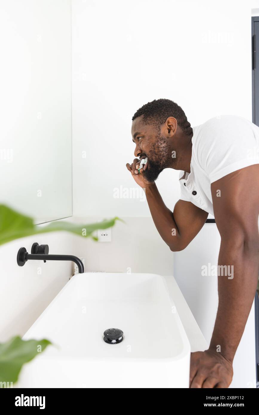 Un giovane afroamericano che spazzola i denti in un bagno moderno con pareti bianche luminose, a casa Foto Stock