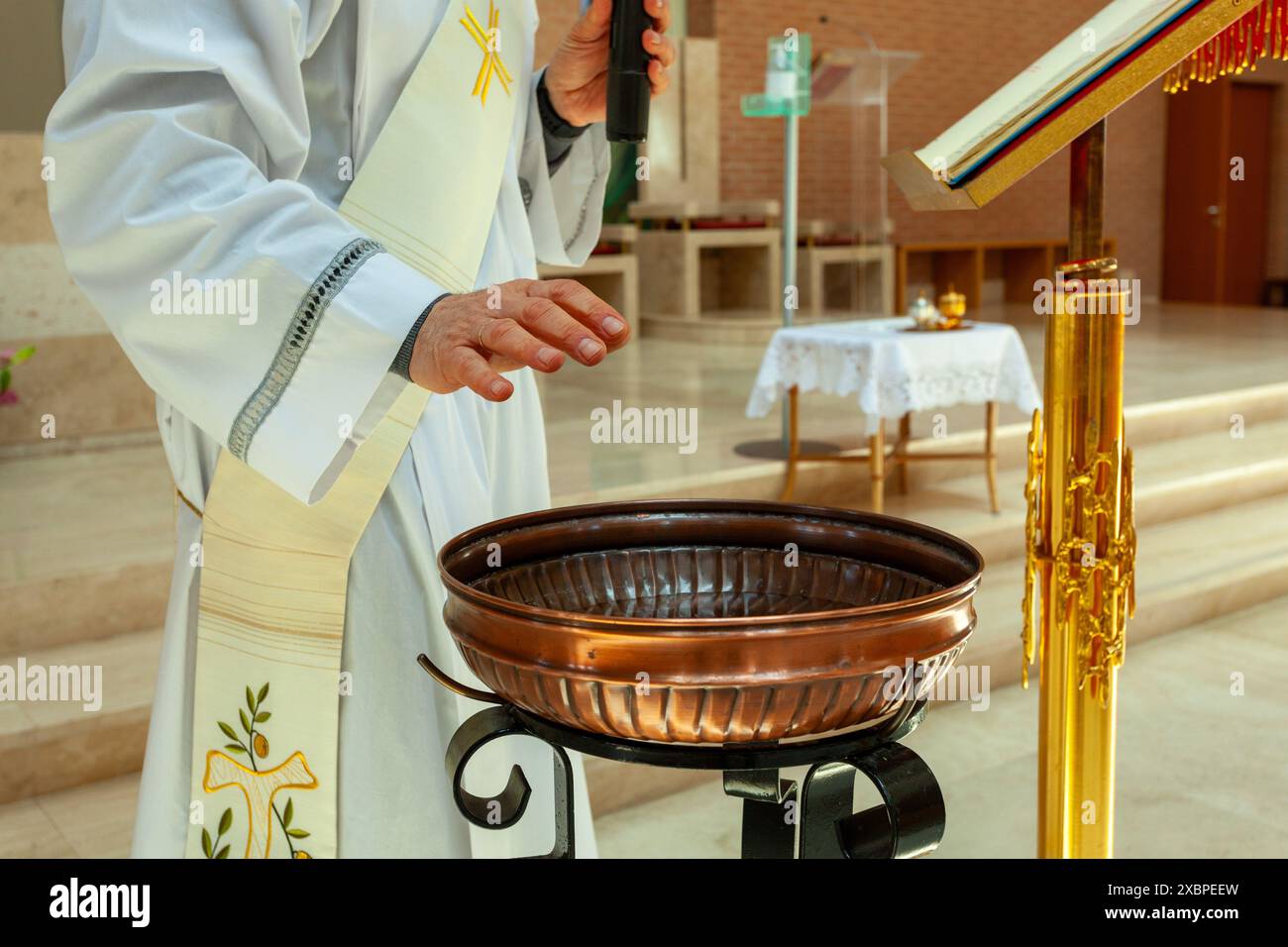 Sacerdote che indossa una veste bianca del clero, in piedi all'altare, tenendo un microfono e preparando il fonte battesimale per la cerimonia Foto Stock