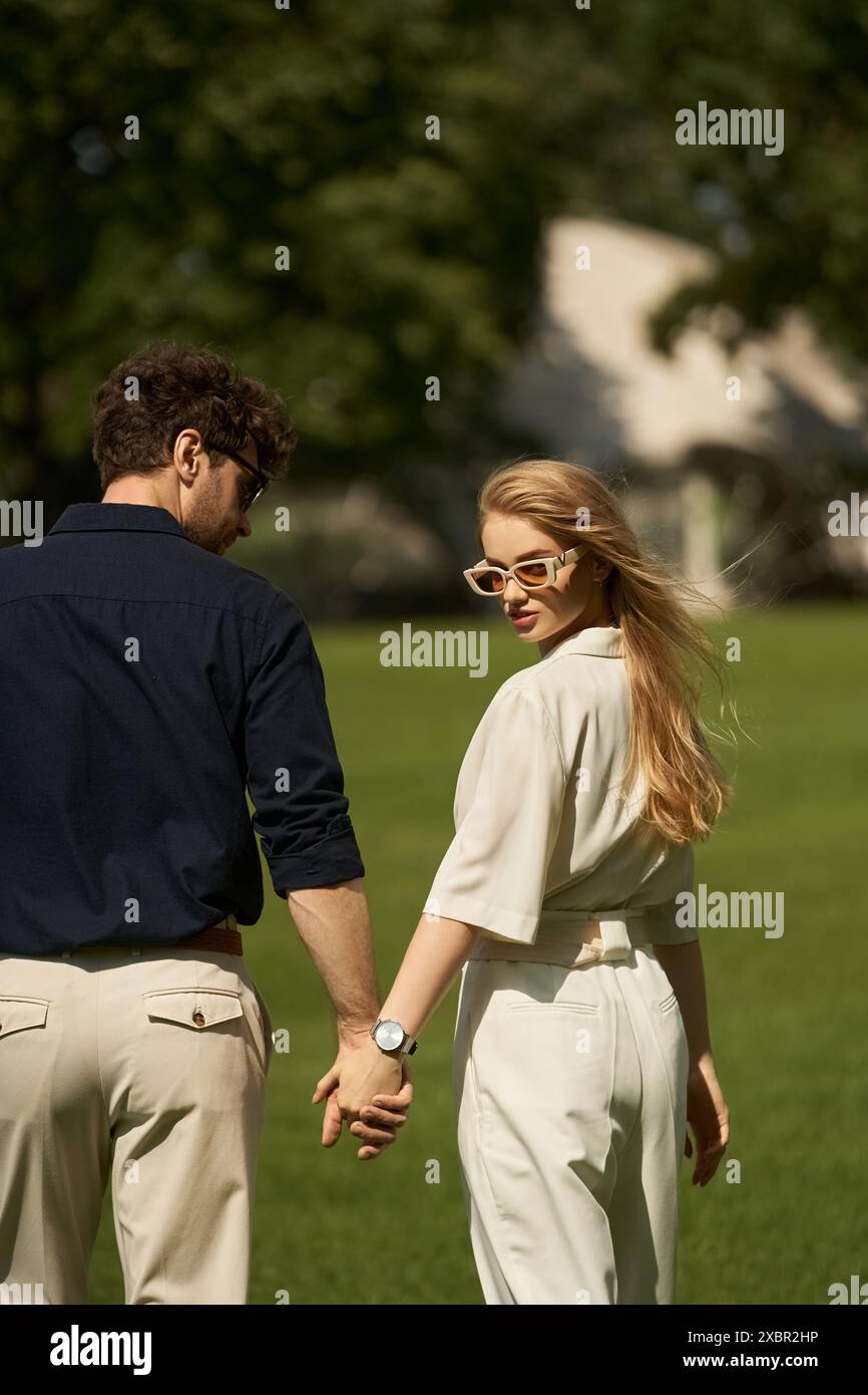 Una giovane coppia elegantemente vestita, tenuta per mano in un parco, circondata da una vegetazione lussureggiante, incarna un senso di eleganza senza tempo. Foto Stock