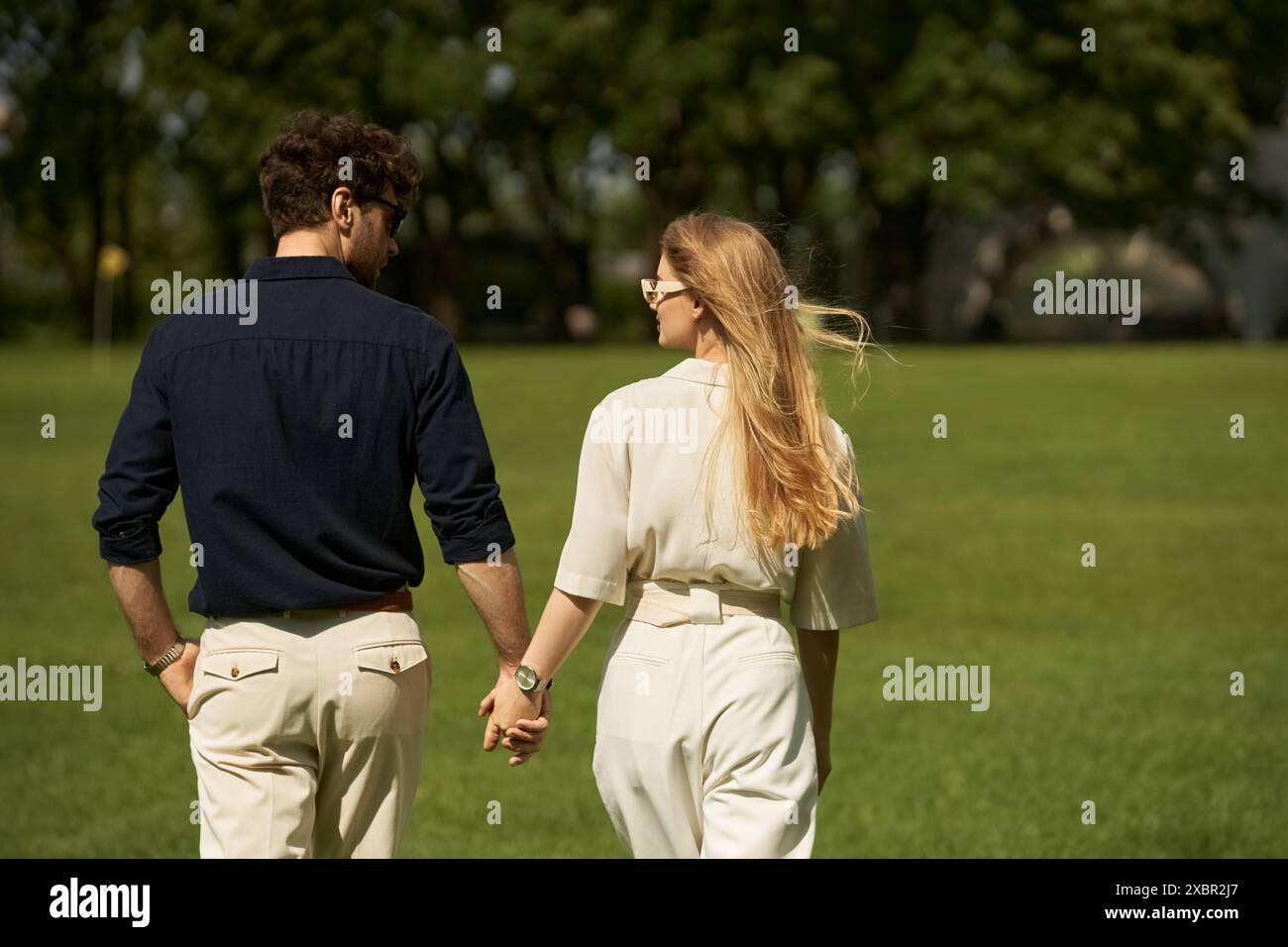 Una bella giovane coppia con un abbigliamento elegante che si tiene per mano mentre cammina attraverso un parco in una giornata di sole. Foto Stock