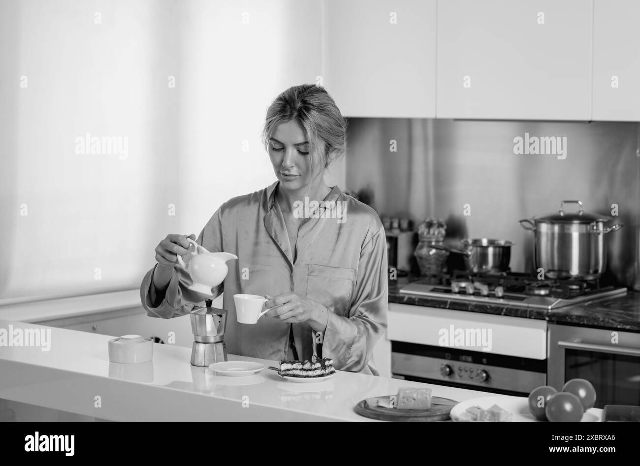 Bella donna che versa il latte nel caffè vicino al tavolo in cucina a casa. Casalinga in cucina al mattino. Foto Stock