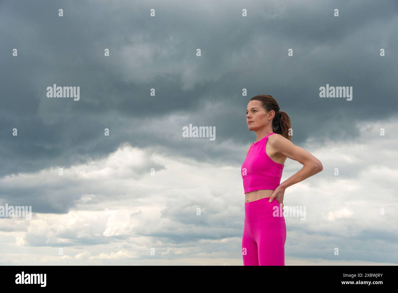 Donna sportiva in piedi che riposa con le mani sui fianchi e indossa abiti sportivi rosa contro un cielo spettacolare. Foto Stock
