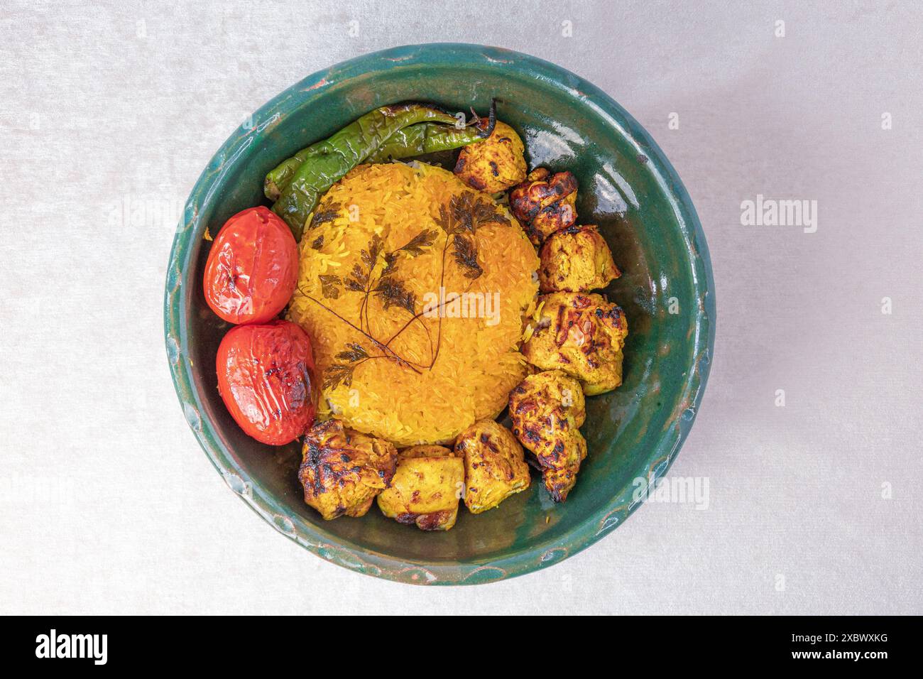 Kebab di pollo persiano (noto anche come jooje kabab) con riso allo zafferano, pomodori grigliati e peperoncini in una ciotola di argilla verde, su sfondo bianco Foto Stock