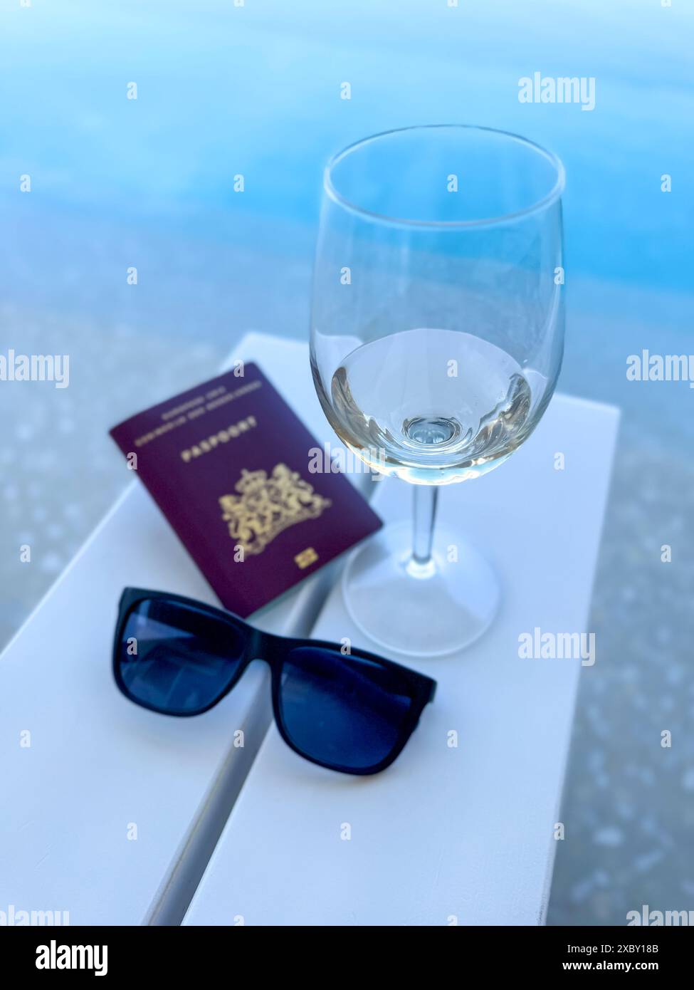 Un'immagine di viaggio con occhiali da sole, vino bianco e un passaporto su un tavolo che si affaccia su una splendida piscina a sfioro che evoca un senso di lusso e di vagabondaggio Foto Stock