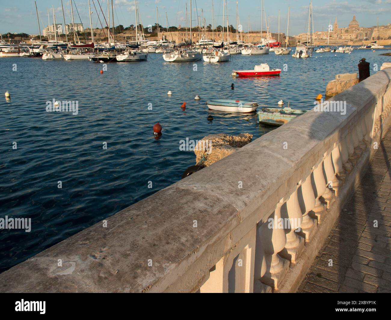 Balaustra in pietra lungo la riva del porto con molte barche in acqua e città sullo sfondo, la valletta, il mediterraneo, malta Foto Stock