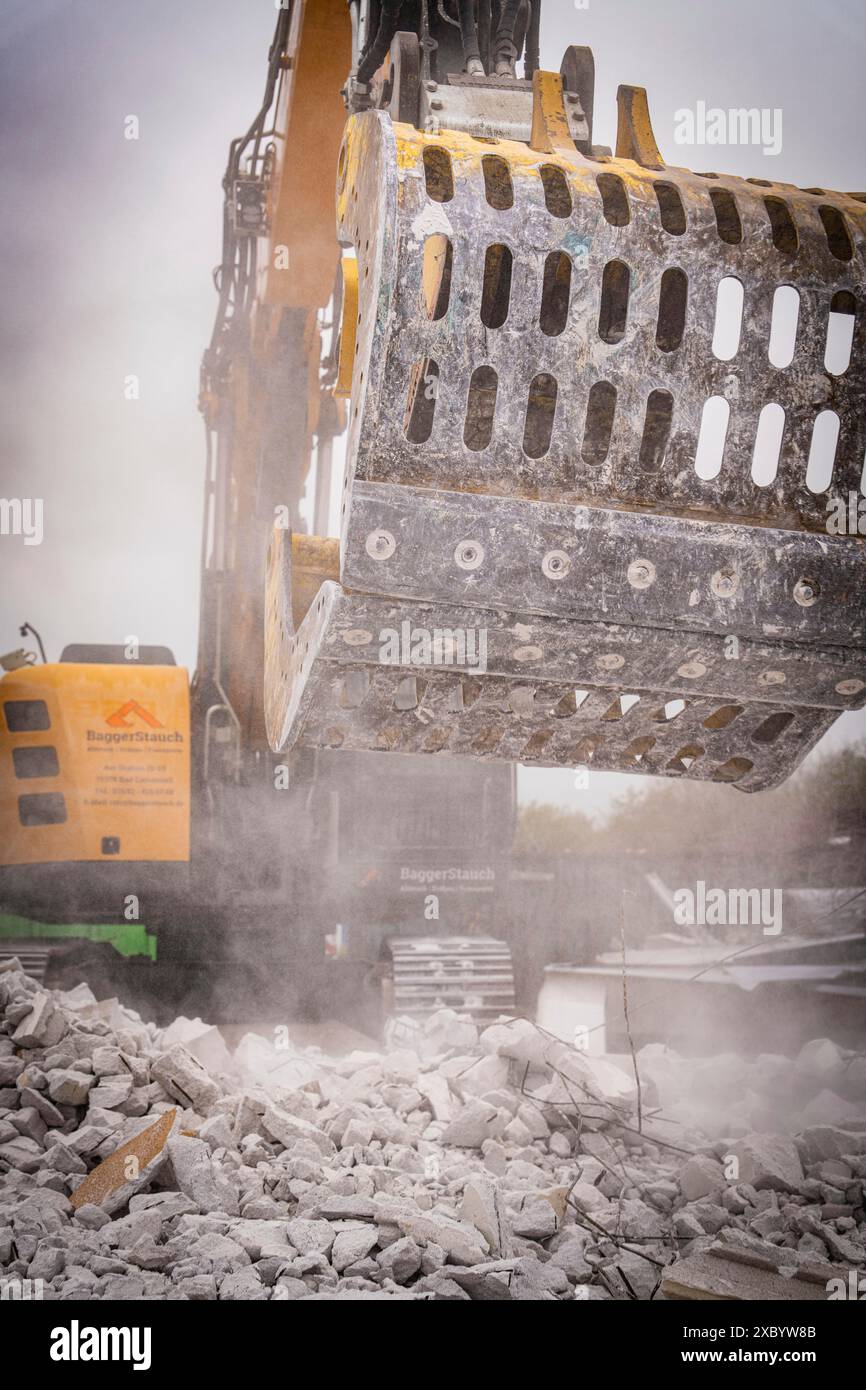 L'escavatore da demolizione con benna metallica frantuma le macerie di calcestruzzo, solleva la polvere in un cantiere, in un cantiere di demolizione, a Saarbruecken, Germania Foto Stock