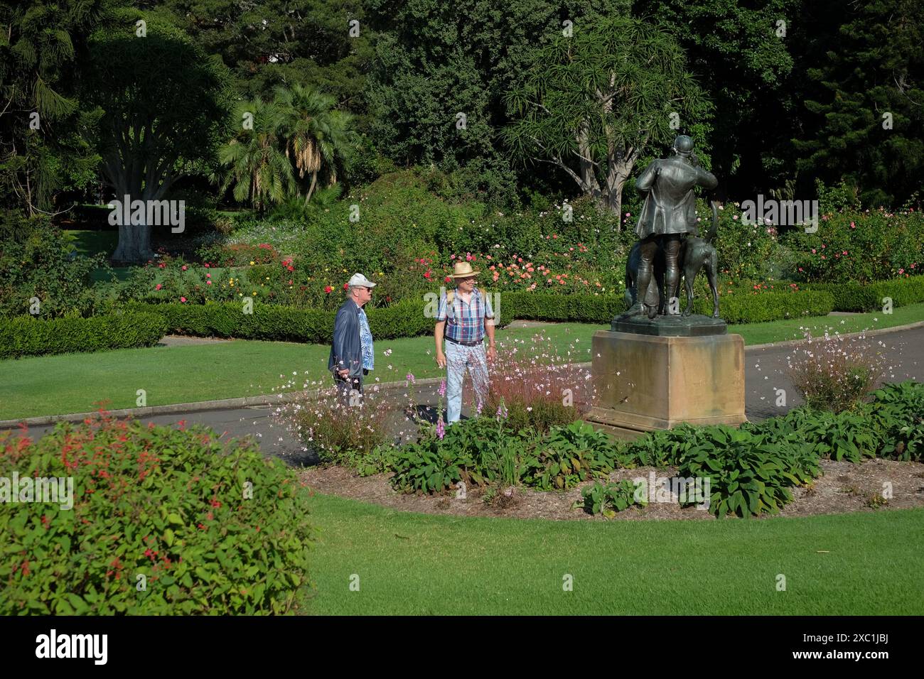 Due uomini osservano la statua di Huntsman e cani nei Royal Botanical Gardens di Sydney con prati verdi e alberi consolidati e piantagioni di confine Foto Stock