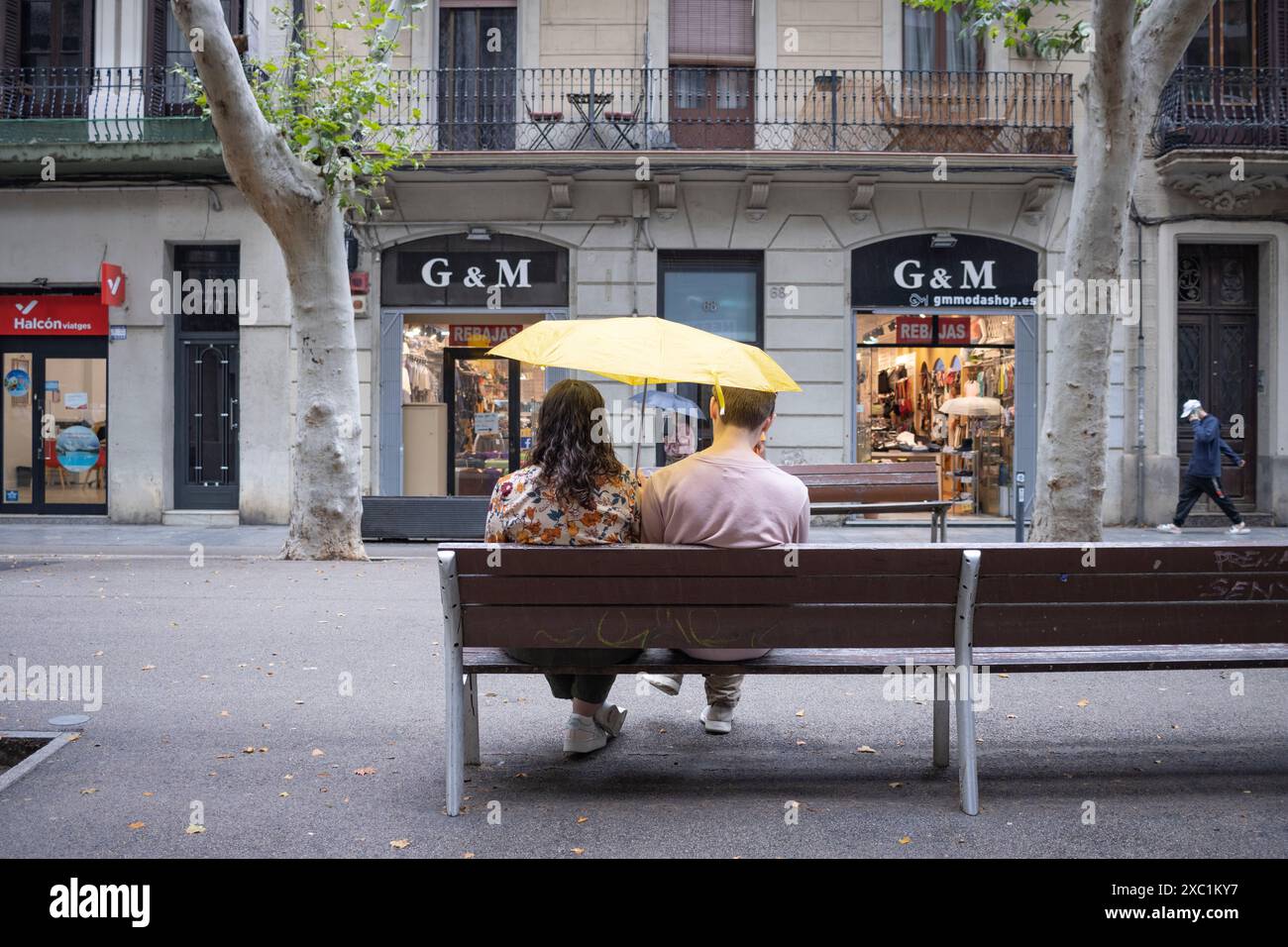 Una giovane coppia seduta su una panchina sotto un ombrello giallo a Rambla del Poblenou, Barcellona, Spagna Foto Stock