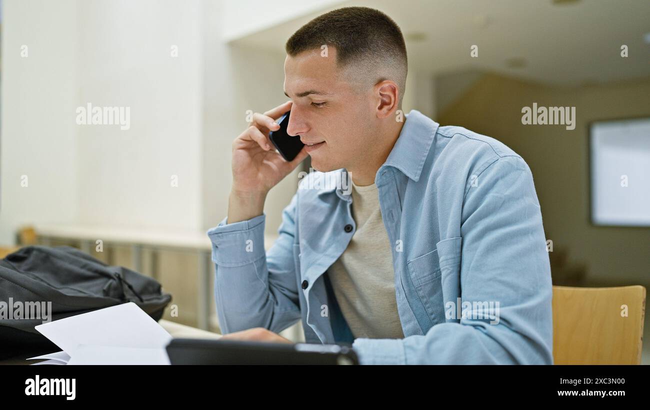 Un giovane fa una telefonata mentre studia con i documenti e un portatile in una biblioteca. Foto Stock