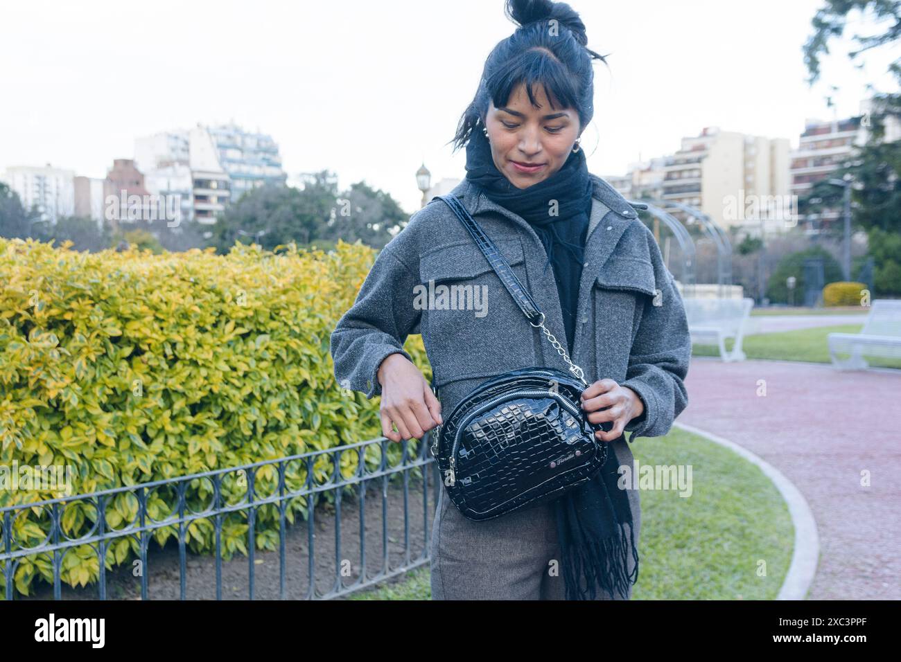 Donna latina nel parco che ara tenendo il portafoglio di pelle aprirlo per prendere qualcosa. Indossa una giacca grigia e una sciarpa, è silenziosa Foto Stock