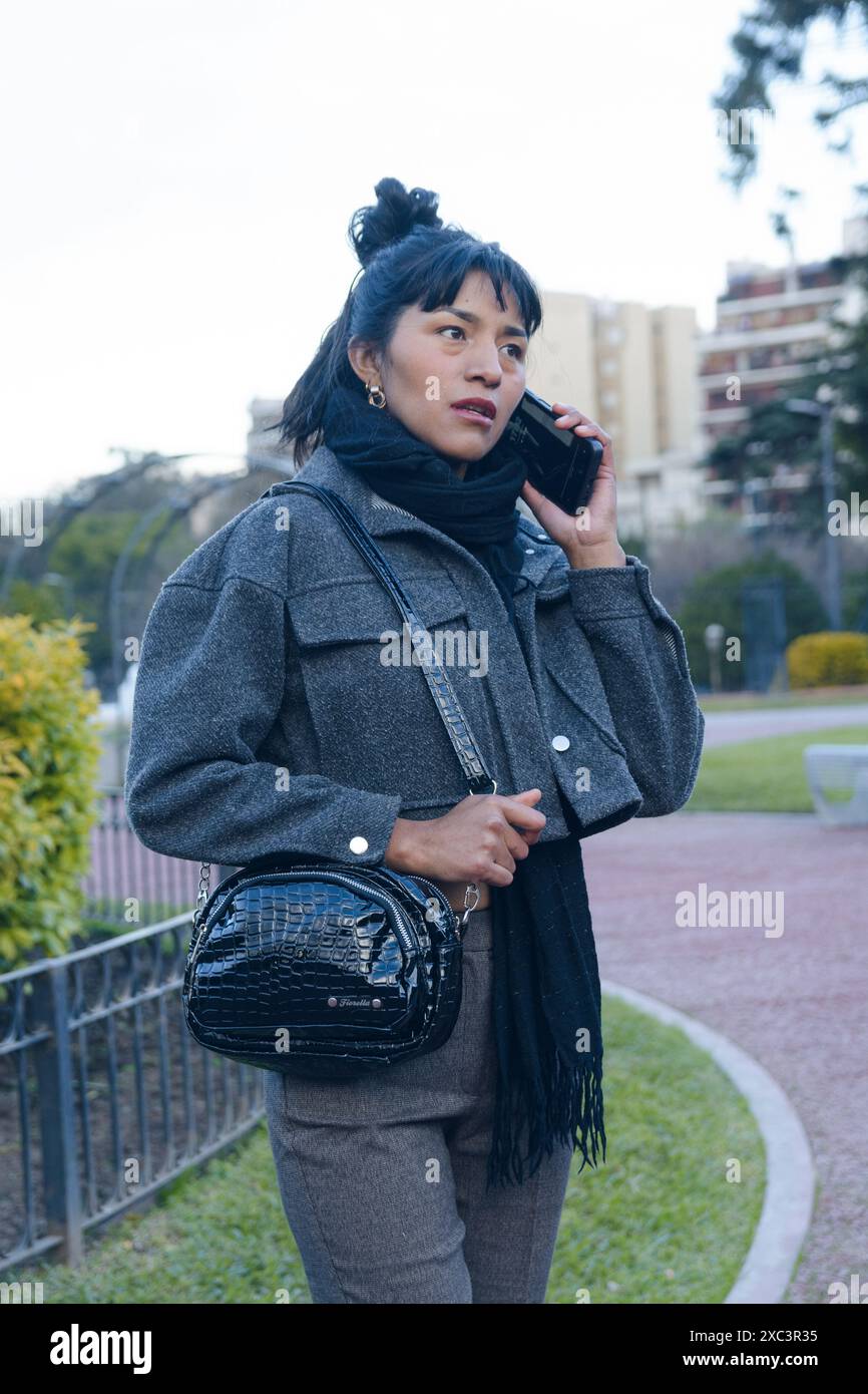 Una donna latina all'aperto sta parlando al suo cellulare mentre tiene una borsa nera. Indossa una giacca grigia e pantaloni neri Foto Stock