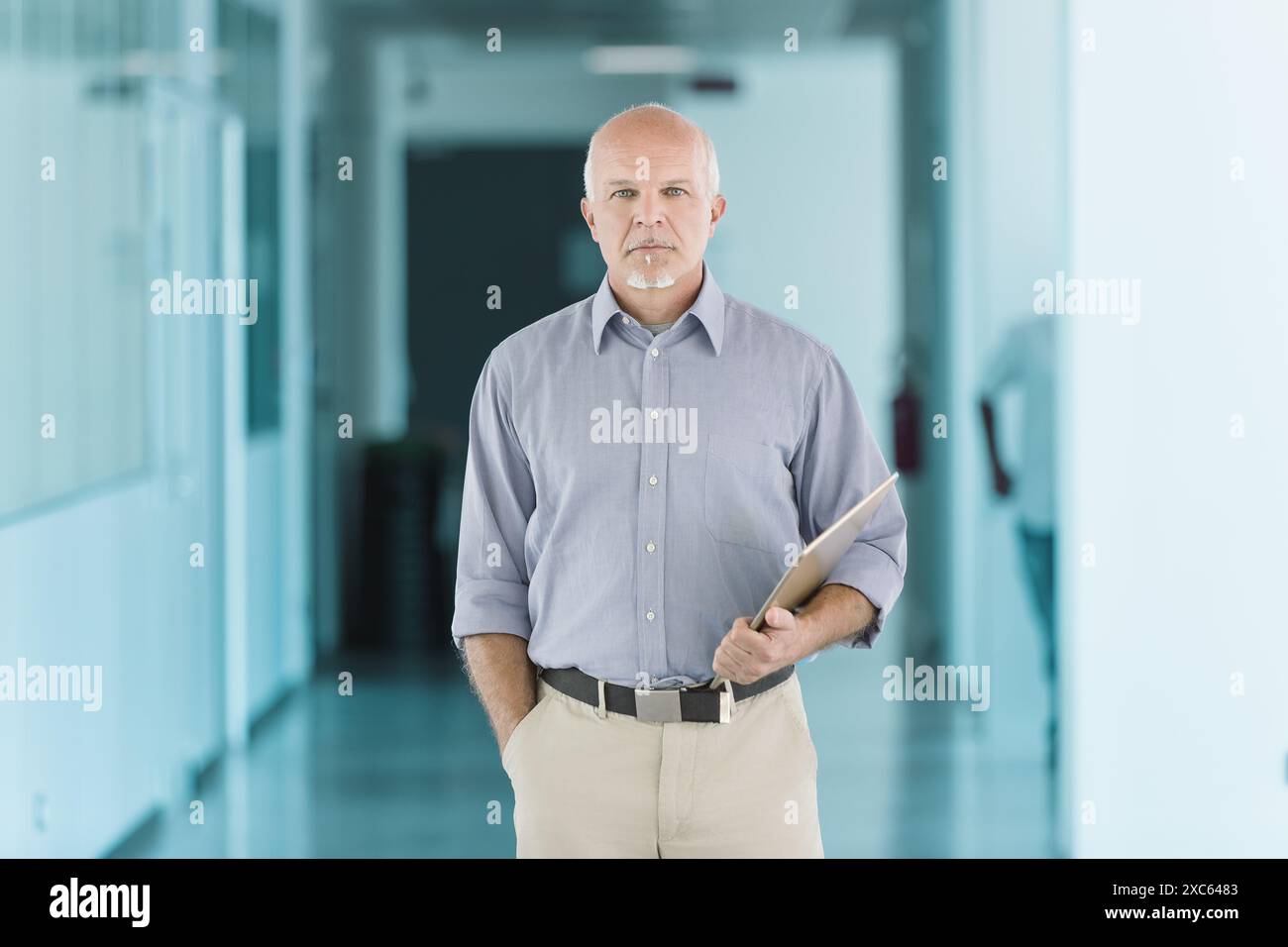 uomo calvo con un pizzetto bianco, con una camicia azzurra e pantaloni beige, si trova in un corridoio con una piegatrice. La sua espressione seria è incorniciata da TH Foto Stock