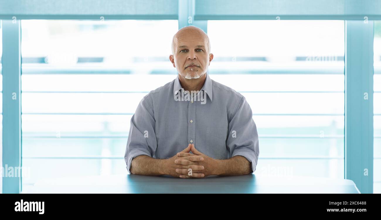 uomo calvo di mezza età con un pizzetto bianco e una camicia azzurra si siede a un tavolo, le mani stringute, guardando direttamente avanti. Lo sfondo luminoso con horiz Foto Stock