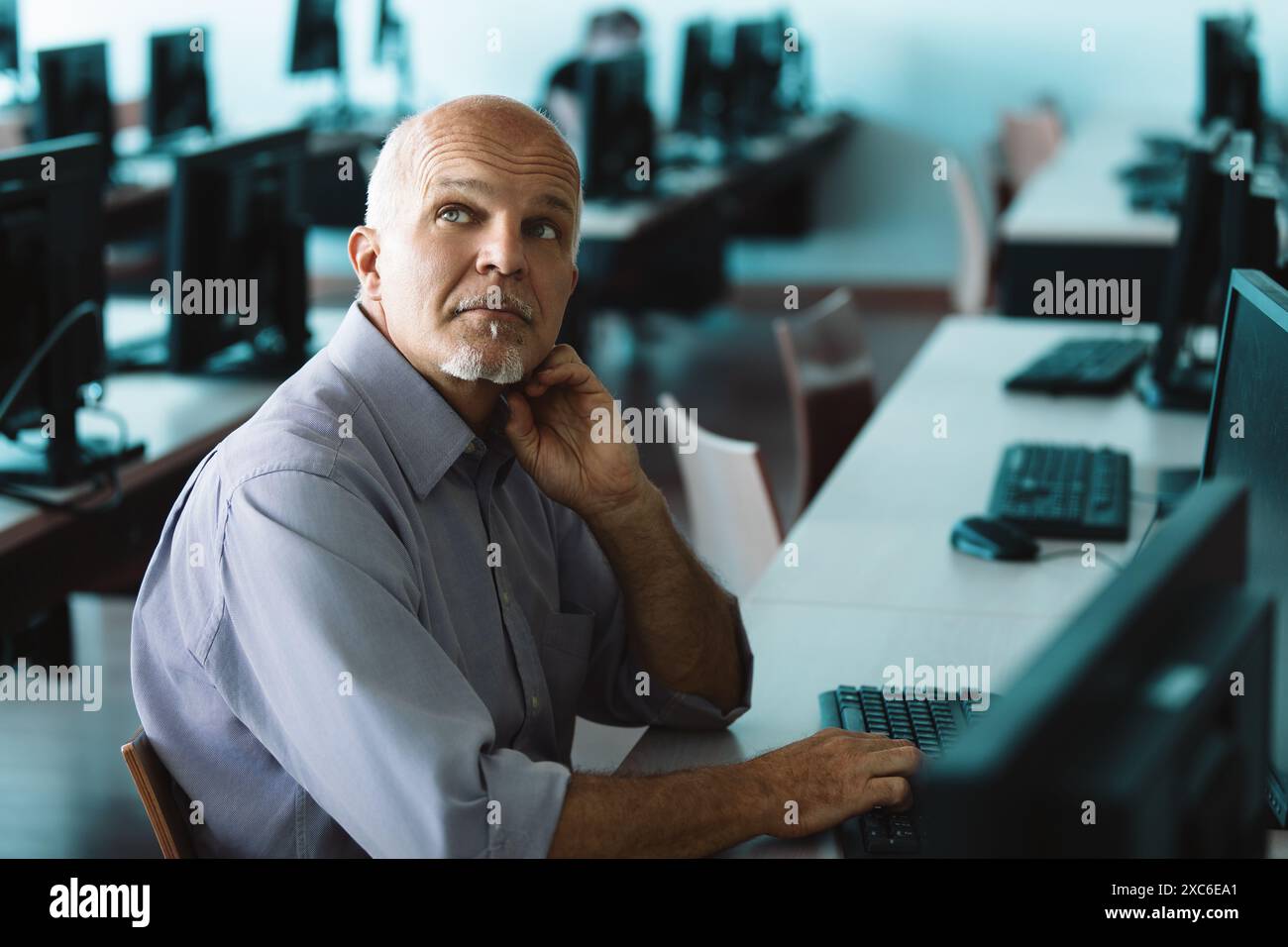 Uomo maturo e concentrato alla scrivania, codifica e risoluzione dei problemi su computer in un ufficio pieno di tecnologia. Programmatore professionista alla ricerca di soluzioni innovative Foto Stock