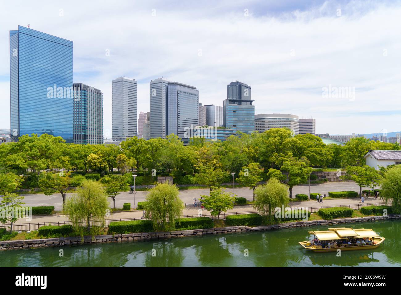 Fiume delimitato da vegetazione lussureggiante, con uno sfondo di grattacieli torreggianti e moderni di Osaka. Una barca gialla in stile tradizionale naviga lungo le acque calme, a. Foto Stock