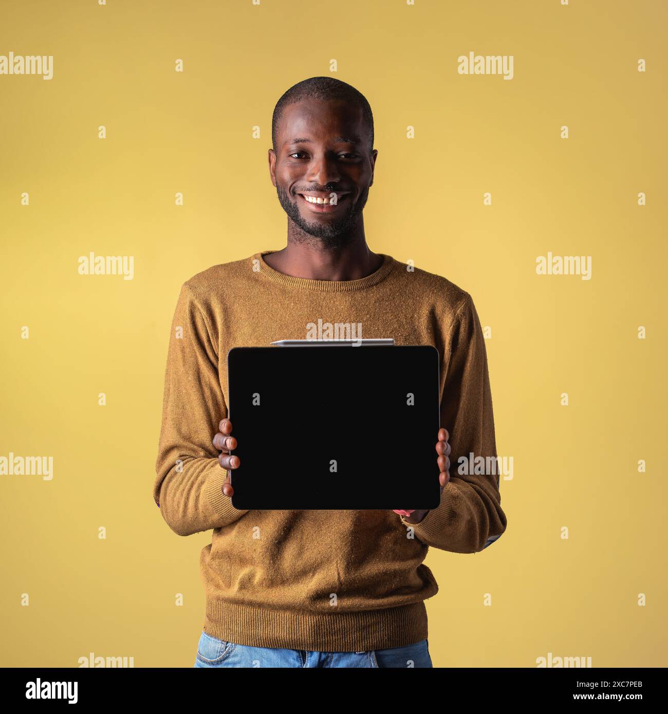 Sorridente giovane afro-americano che tiene in mano un tablet, presenta il dispositivo su uno sfondo giallo - uomo felice che mostra lo schermo digitale - moderno Technolo Foto Stock