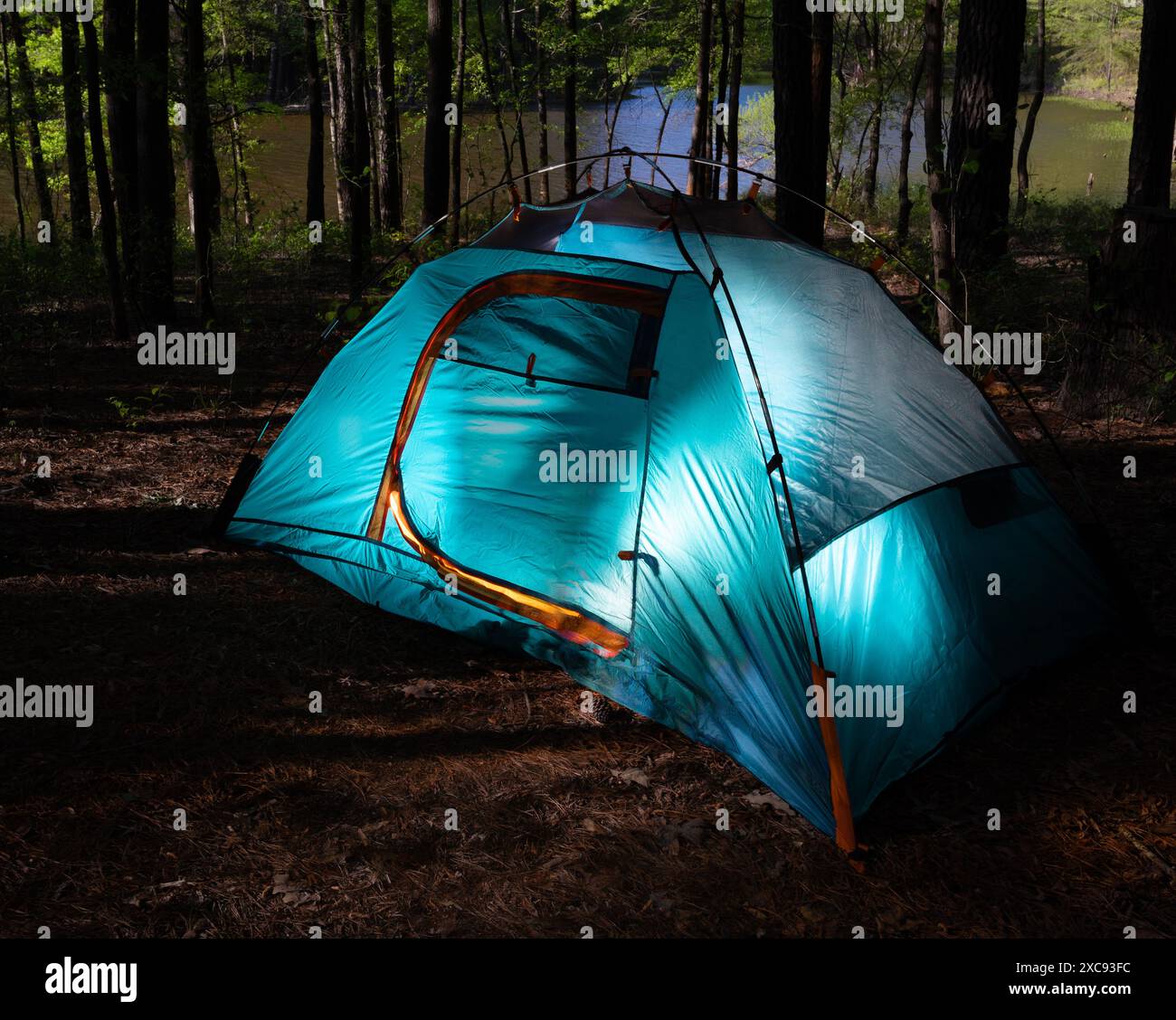 Tenda da da zaino azzurra illuminata in una fitta foresta con dietro il lago, proprio mentre si mostra un po' di luce solare, rivelando l'attrazione di un semplice ou Foto Stock