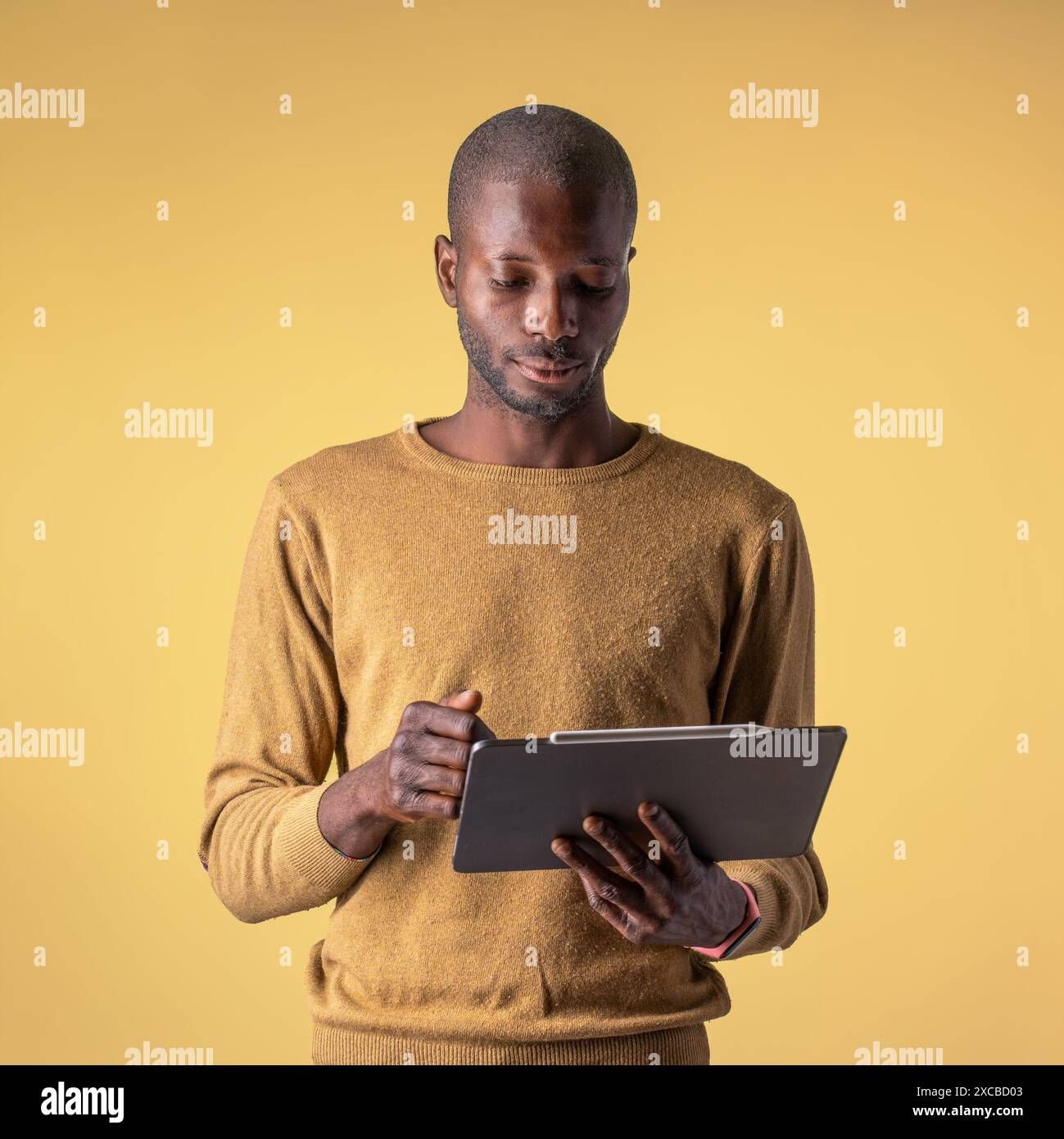 Un giovane afroamericano che usa un tablet, in piedi casualmente su uno sfondo giallo - uso tecnologico informale - atmosfera moderna e focalizzata, vita digitale Foto Stock