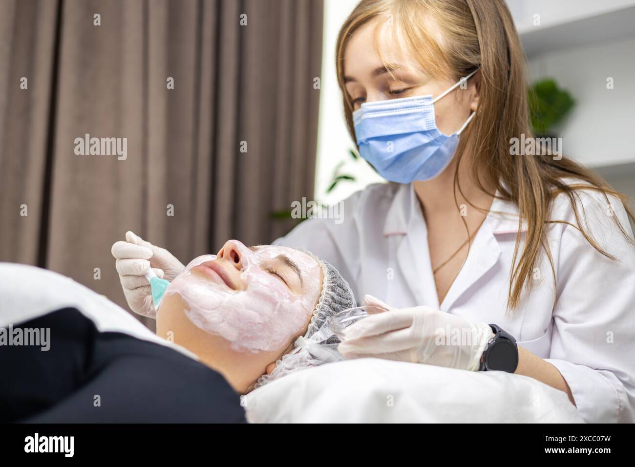 Un ritratto di profilo laterale di un giovane cosmetologo che indossa guanti bianchi e maschera chirurgica, applicando una maschera facciale con una spatola sul viso di un cliente. Foto Stock