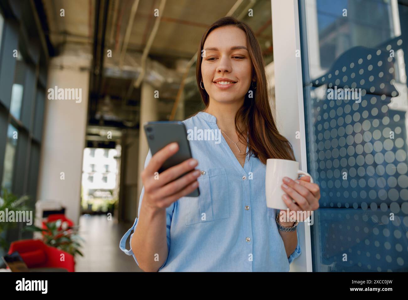 Una donna sembra felice di sfogliare il suo smartphone tenendo una tazza di caffè in un ufficio alla moda Foto Stock