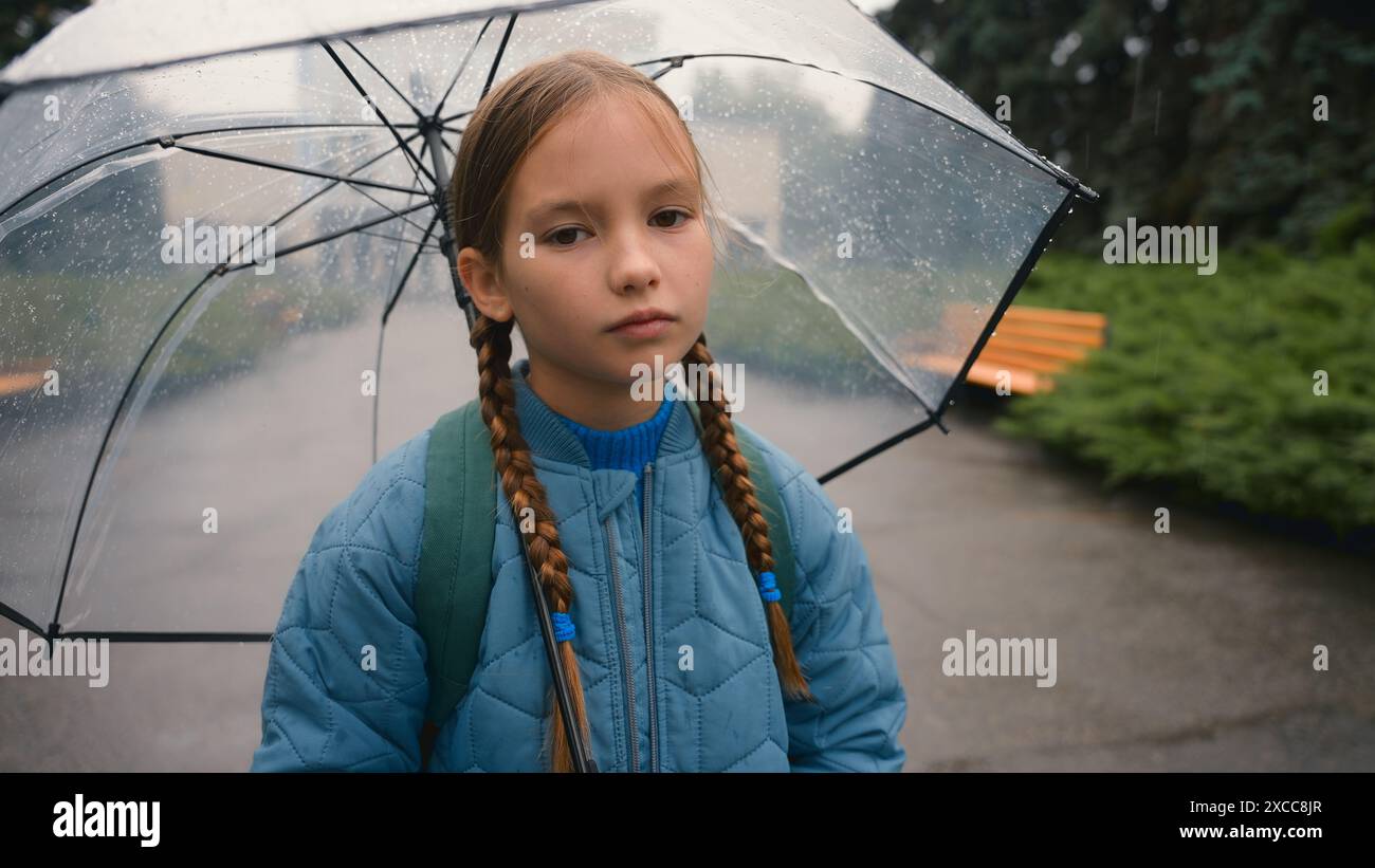 Bambina stanca studentessa bambino che guarda la macchina fotografica annoiato maltempo ombrello pioggia parco cittadino all'aperto fatica bagnato viso figlia bambina Foto Stock