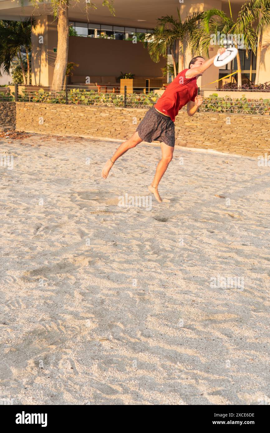 Un giovane con una camicia rossa e pantaloncini saltano per prendere un frisbee su una spiaggia sabbiosa, con uno sfondo di verde e pareti di pietra, durante il tramonto. Foto Stock