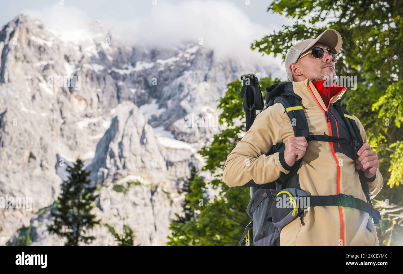 Un uomo con uno zaino in piedi che guarda le vette innevate delle Alpi. Indossa un berretto, occhiali da sole e una giacca abbronzante. Foto Stock