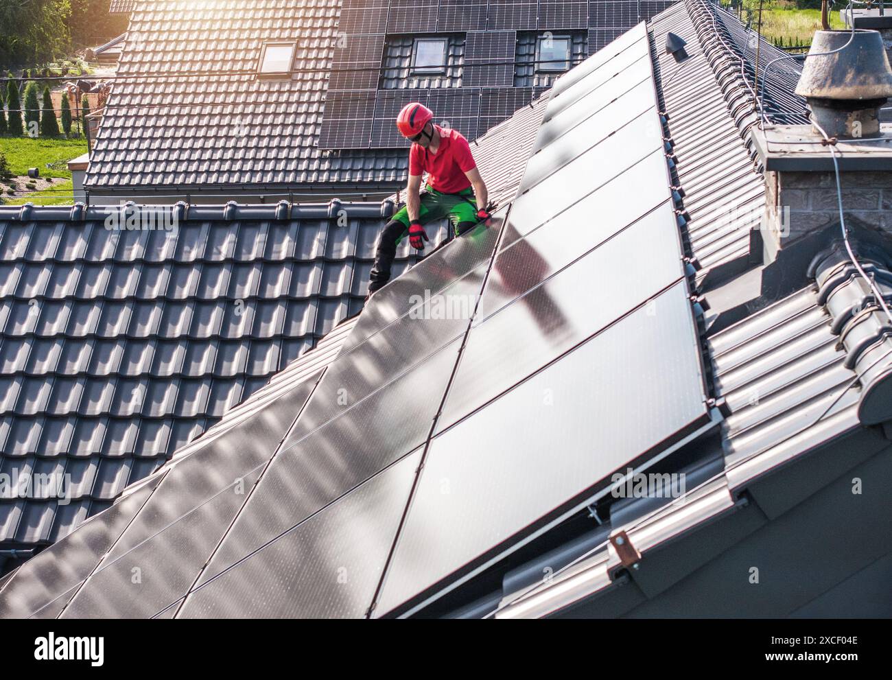 Un lavoratore che indossa un elmetto rosso e pantaloni verdi installa pannelli solari su un tetto residenziale. Il tetto è piastrellato e i pannelli sono disposti in file. Foto Stock