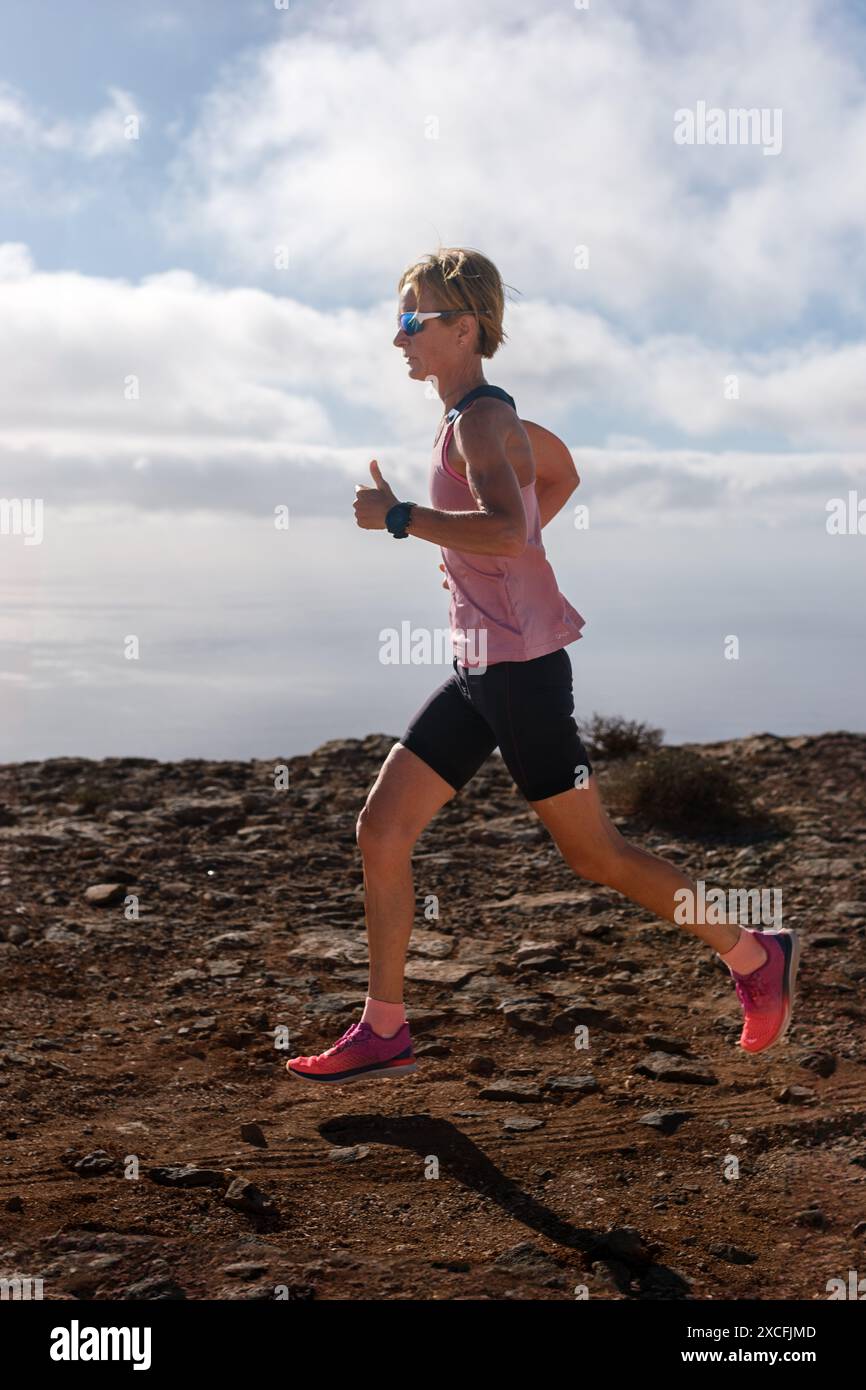 Percorrendo un paesaggio arido e roccioso, una donna con canotta rosa e shorts neri rimane concentrata sul suo percorso, la sua forma atletica ha sagomato agains Foto Stock Percorrendo un paesaggio arido e roccioso, una donna con canotta rosa e shorts neri rimane concentrata sul suo percorso, la sua forma atletica ha sagomato agains Foto Stock