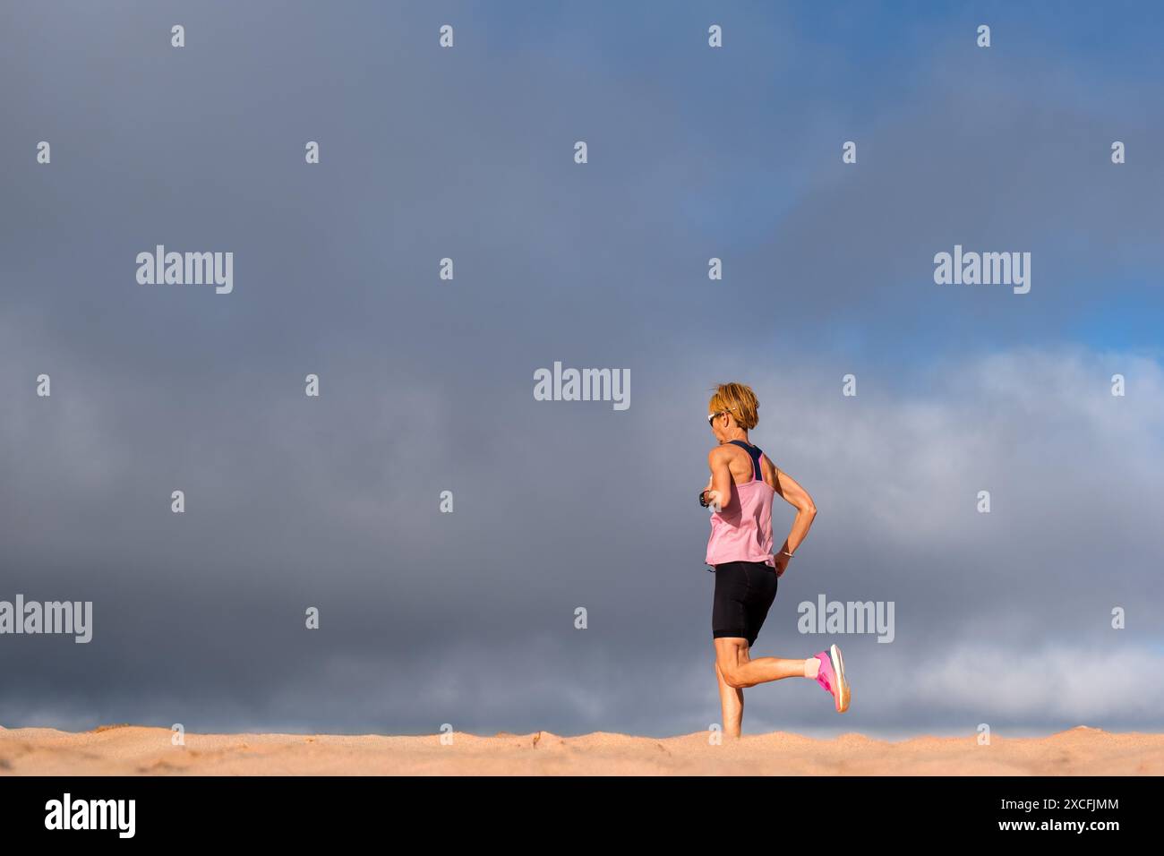 Una donna corre all'aperto indossando abbigliamento fitness sotto un cielo blu nuvoloso, incarnando salute, fitness e determinazione. Foto Stock