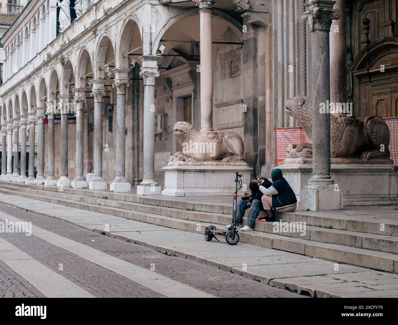 Cremona, Italia - 10 giugno 2024 due persone fanno una pausa sui gradini di un edificio storico con uno scooter elettrico parcheggiato accanto a loro Foto Stock