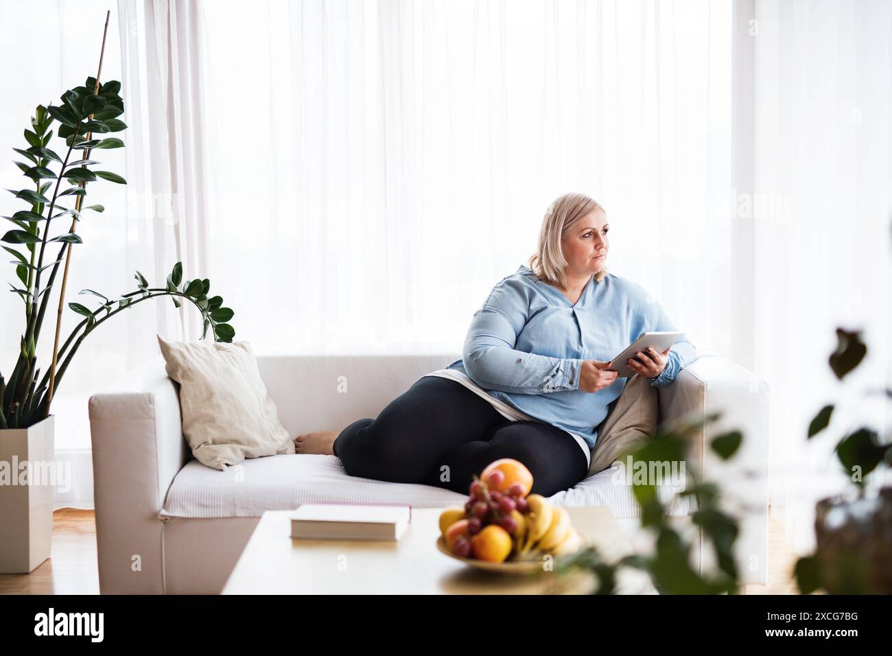 Ritratto di una bella giovane donna in sovrappeso a casa, che guarda programmi tv su tablet, si siede sul divano in soggiorno e si gode una tranquilla giornata del fine settimana. Beaut Foto Stock
