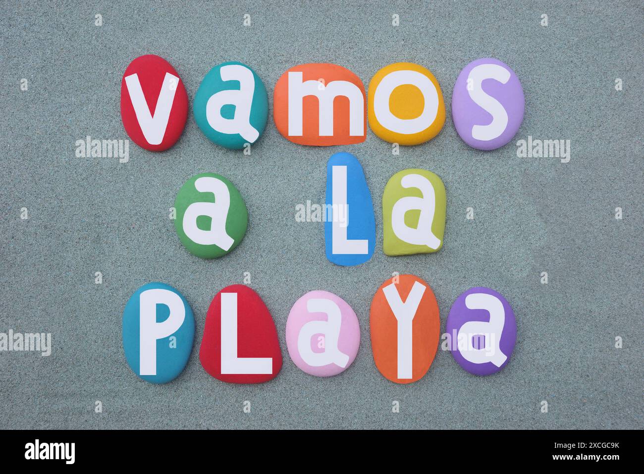 Vamos a la playa, andiamo in spiaggia, creativo slogan estivo spagnolo composto da lettere in pietra dipinte a mano multicolore su sabbia verde Foto Stock