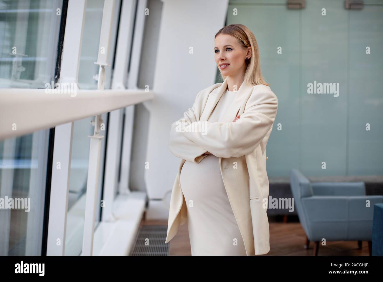 Bella e sicura donna bionda incinta in un abbigliamento elegante e alla moda, con un abito bianco e un blazer, in piedi con le braccia incrociate in un'azienda Foto Stock