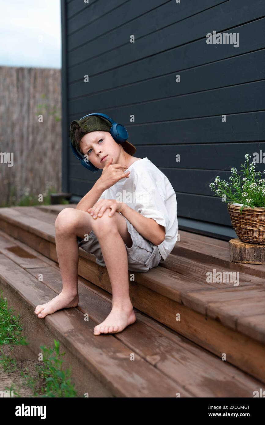 Simpatico ragazzo preadolescente con le cuffie che ascolta la musica su una terrazza in legno all'aperto Foto Stock