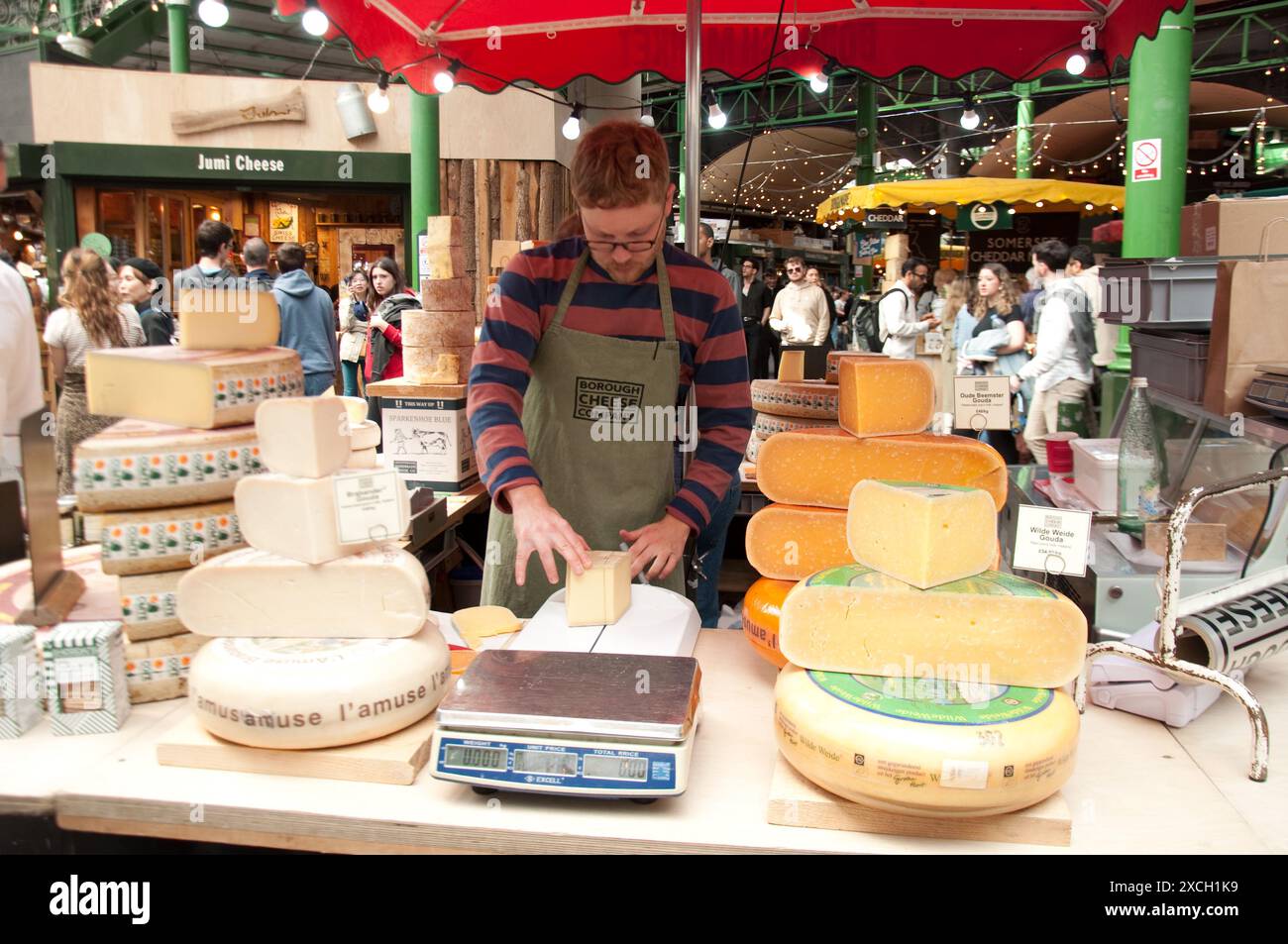 Cheese stall, Borough Market, Bermondsey, Southwark, Londra, Regno Unito - formaggi francesi e olandesi - Gouda; man affettare porzioni di formaggio; molte persone in backg Foto Stock