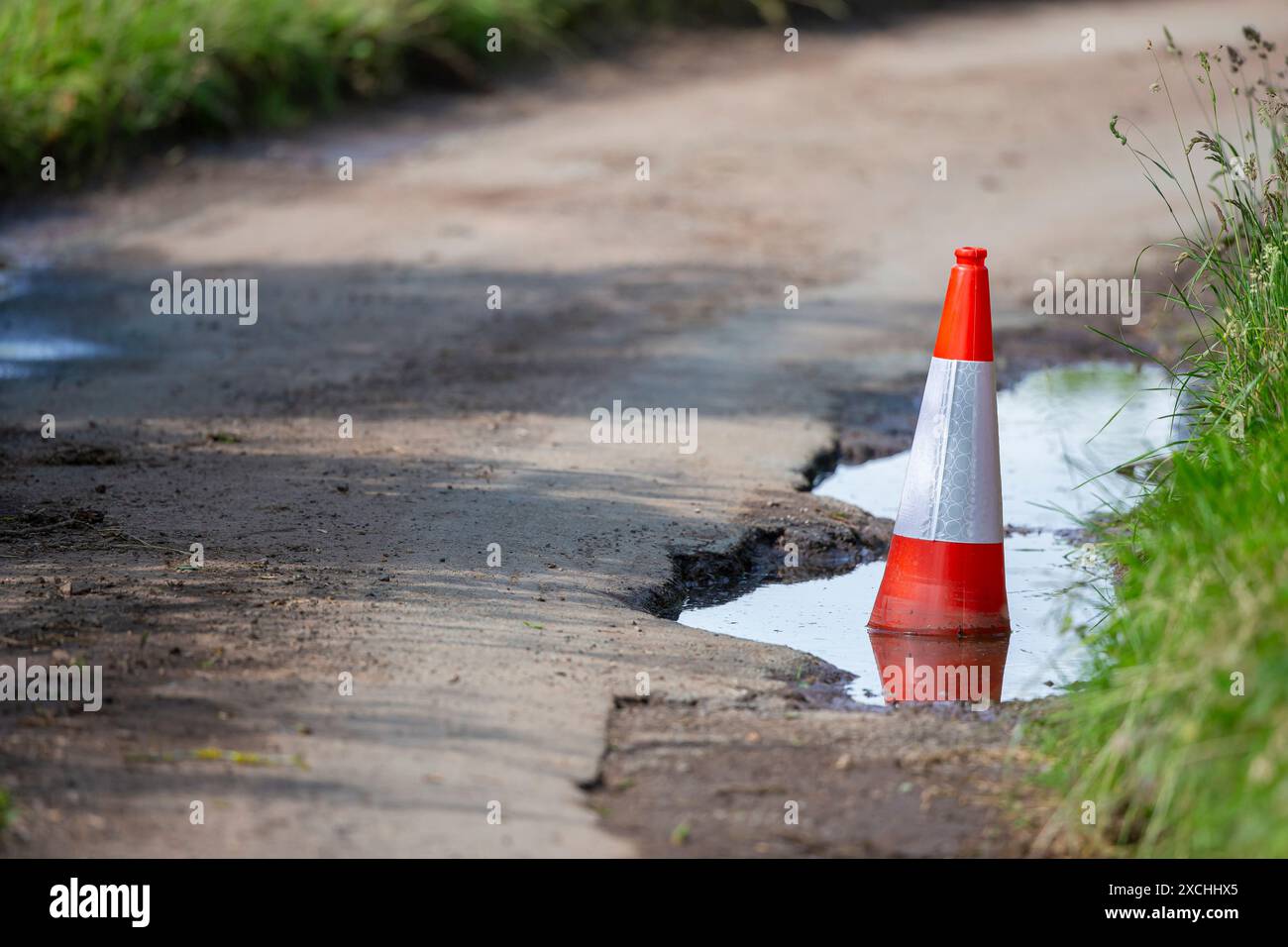 Buca profonda in una corsia di campagna piena d'acqua e segnata da un cono stradale. Foto Stock