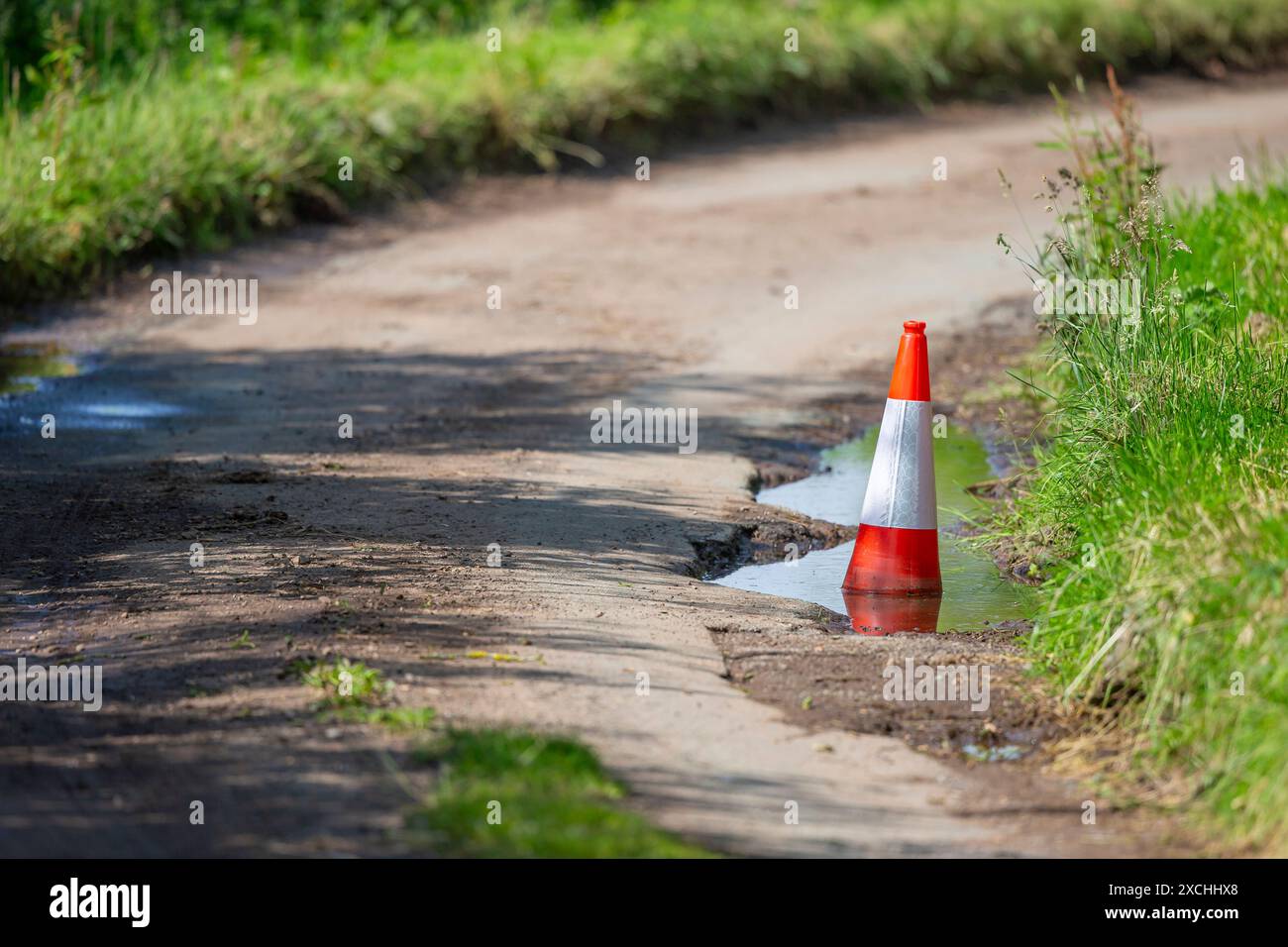 Buca profonda in una corsia di campagna piena d'acqua e segnata da un cono stradale. Foto Stock