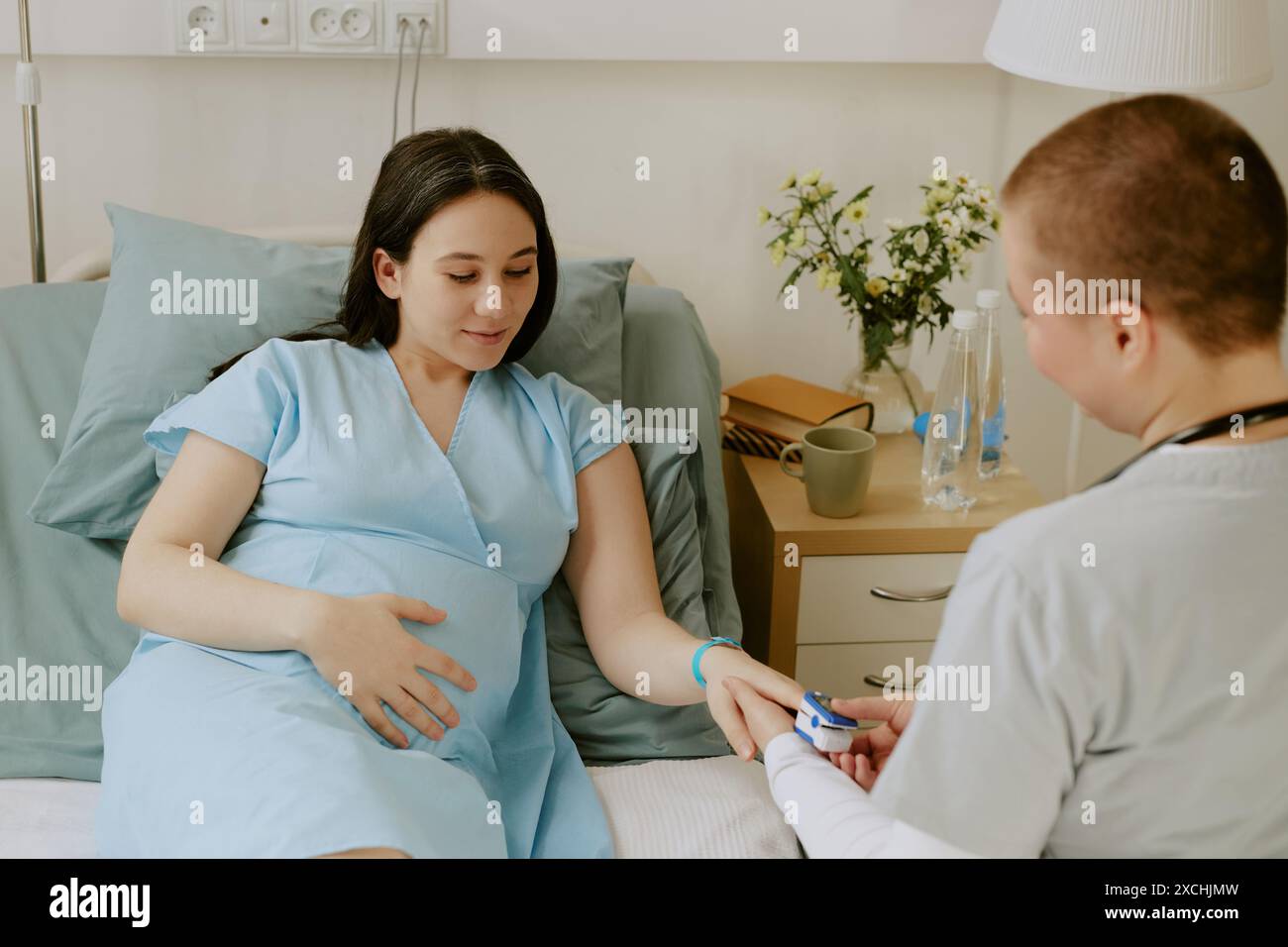 Spalla di una donna incinta sdraiata sul letto e sorridente leggermente mentre il medico controlla la sua saturazione Foto Stock