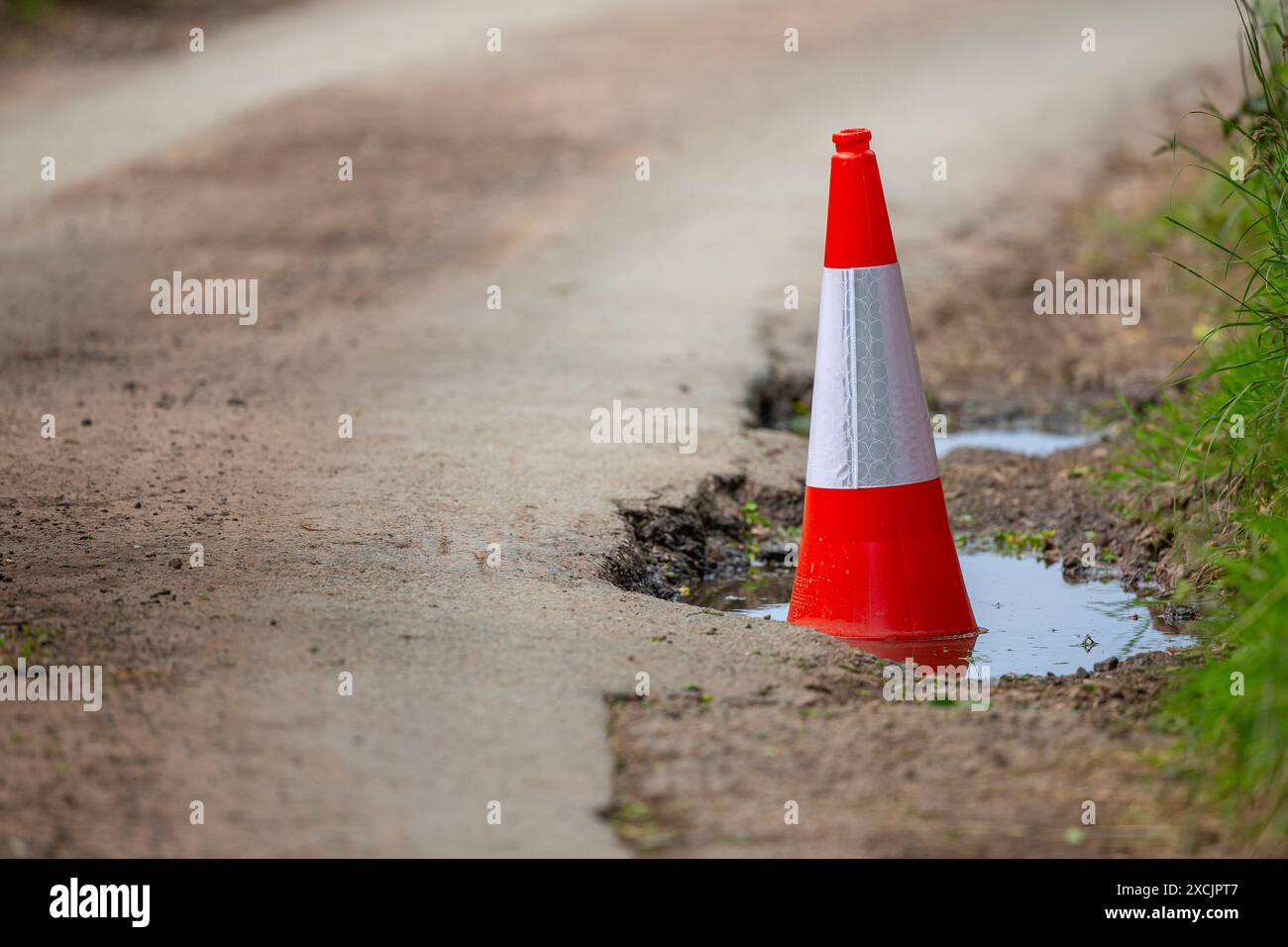 Buca profonda in una corsia di campagna del Regno Unito piena d'acqua e segnata da un cono rosso e bianco della strada. Sicurezza stradale britannica. Foto Stock