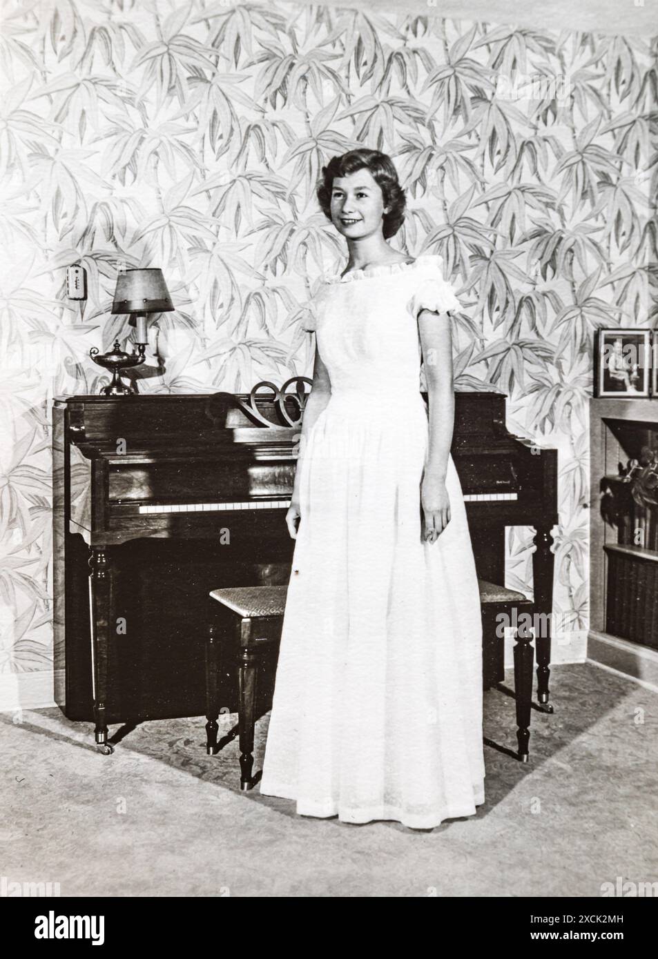 Giovane donna attraente che indossa un abito da ballo per la laurea scolastica in casa con un pianoforte preso nei primi anni '1950 negli Stati Uniti Foto Stock