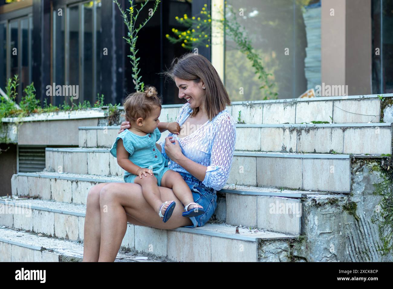 Una giovane madre e il suo bambino sono seduti sui gradini di un edificio in rovina. Il bambino chiede del latte. Foto Stock