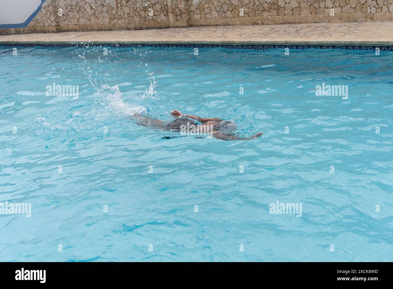 Donna che nuota con il seno in una piscina azzurra, indossa un berretto e un costume da bagno nero. Foto Stock