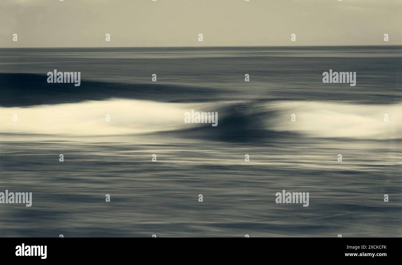 Una tranquilla scena di un'onda dolce che si infrangono su una spiaggia sabbiosa mentre il sole tramonta, che proietta un caldo bagliore sull'acqua e sulla sabbia. L'acqua è un turchese vibrante Foto Stock